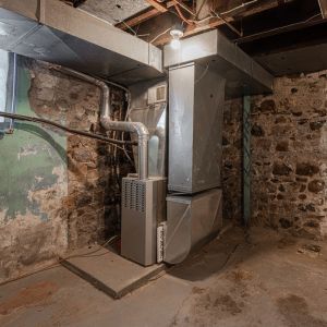 Old gas furnace system in a basement with metal ductwork and stone walls – example of a unit ready for replacement
