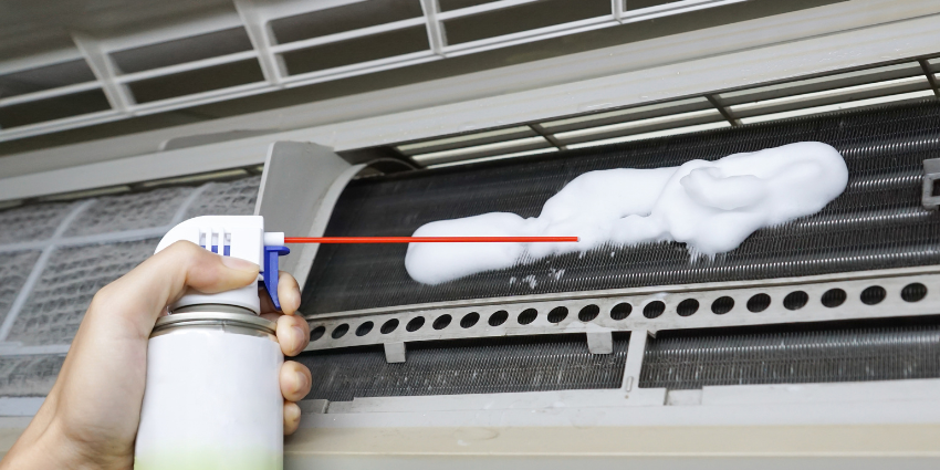 Gloved hand spraying foaming coil cleaner onto the indoor evaporator coil of a Goodman air conditioner.